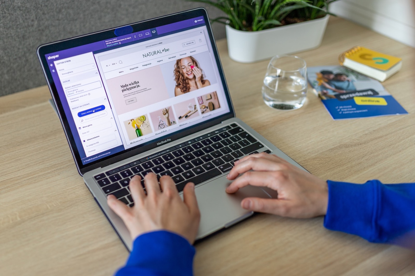 Hands typing on a MacBook Pro displaying an e-commerce website with "NATURAL ME" and beauty products.